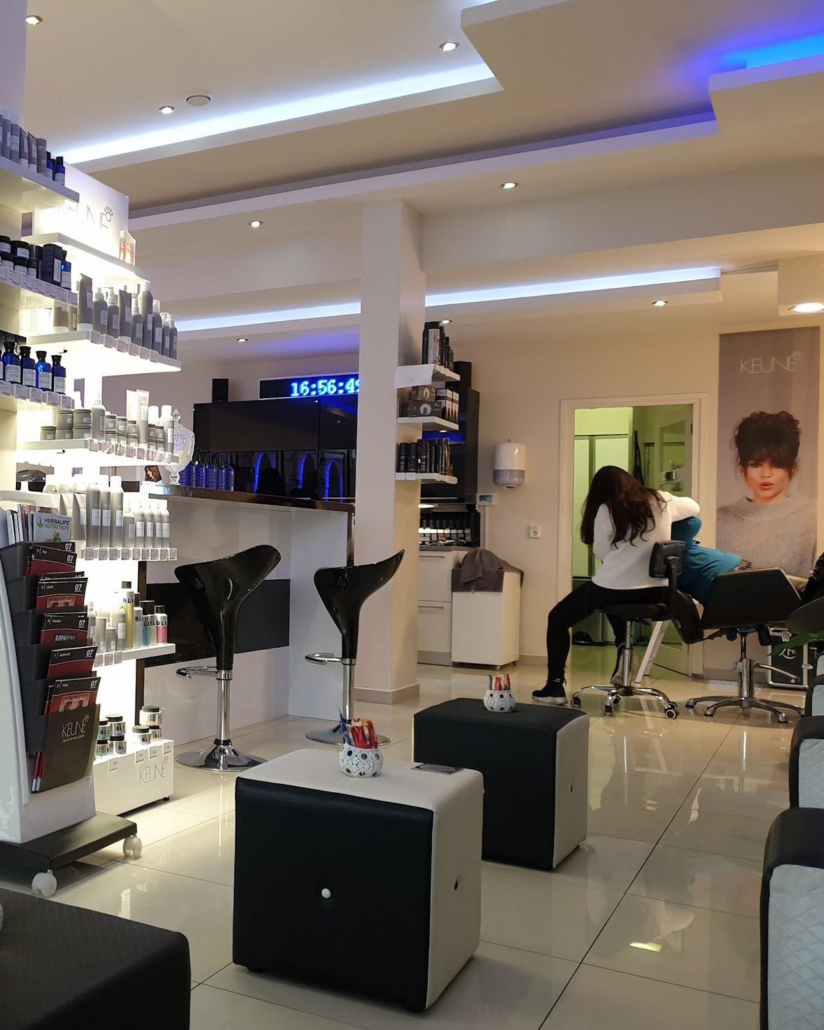 De salon met schappen vol haarproducten, de zwarte barkrukken en achterin een kapster aan het werk bij een klant